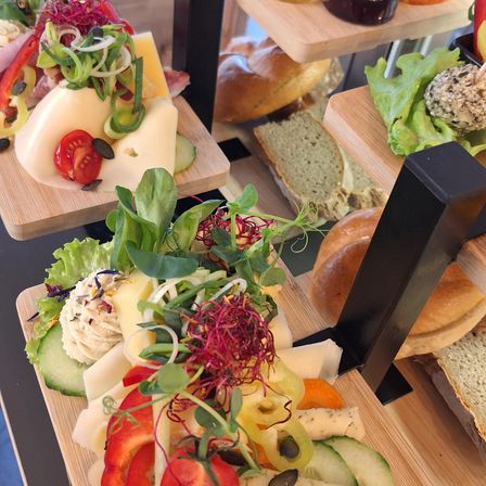 Eine Auswahl an Gourmet-Lebensmitteln auf Holztabletts, einschließlich Käse, Tomaten und Oliven, präsentiert auf einem schwarzen Metallständer.