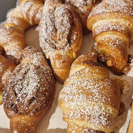 Eine Auswahl an frisch gebackenen Gebäckstücken, darunter Croissants und Zimtschnecken, bestreut mit Puderzucker, auf einer weißen Oberfläche angeordnet.