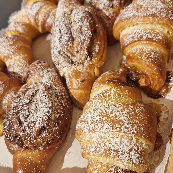 Eine Auswahl an frisch gebackenen Gebäckstücken, darunter Croissants und Zimtschnecken, bestreut mit Puderzucker, auf einer weißen Oberfläche angeordnet.