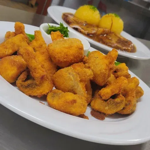 Ein Teller mit knusprigen frittierten Speisen, möglicherweise Tintenfischringe, wird mit einer Schüssel grüner Sauce serviert. Im Hintergrund steht ein weiterer Teller mit Fleisch und Kartoffeln.