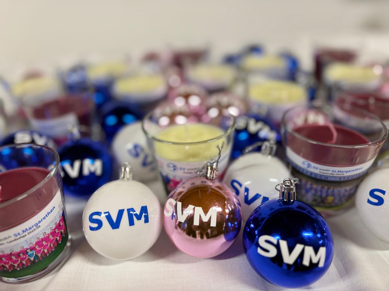 Verschiedene farbige Kugeln mit den Buchstaben SVM darauf sind ausgestellt. Sie befinden sich neben einigen Kerzen in verschiedenen Farben. Die Kerzen sind in Glasbechern.