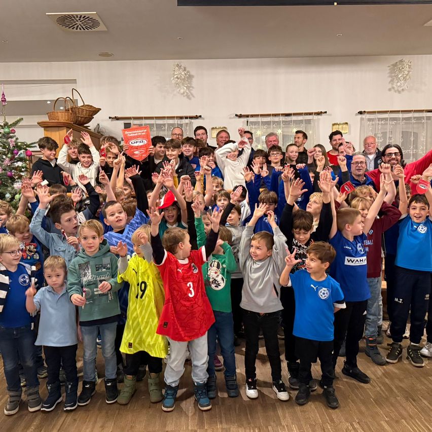 Eine Gruppe von Kindern und Erwachsenen versammelt sich für ein Foto, trägt festliche Kleidung und jubelt, mit einem Weihnachtsbaum im Hintergrund.