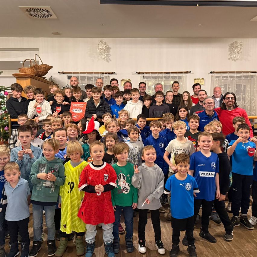 Eine Gruppe von Kindern und Erwachsenen steht für ein Foto zusammen. Die Kinder tragen Sporttrikots und Turnschuhe. Dahinter versammeln sich Erwachsene und Kinder in der Nähe eines Weihnachtsbaums und eines Banners.