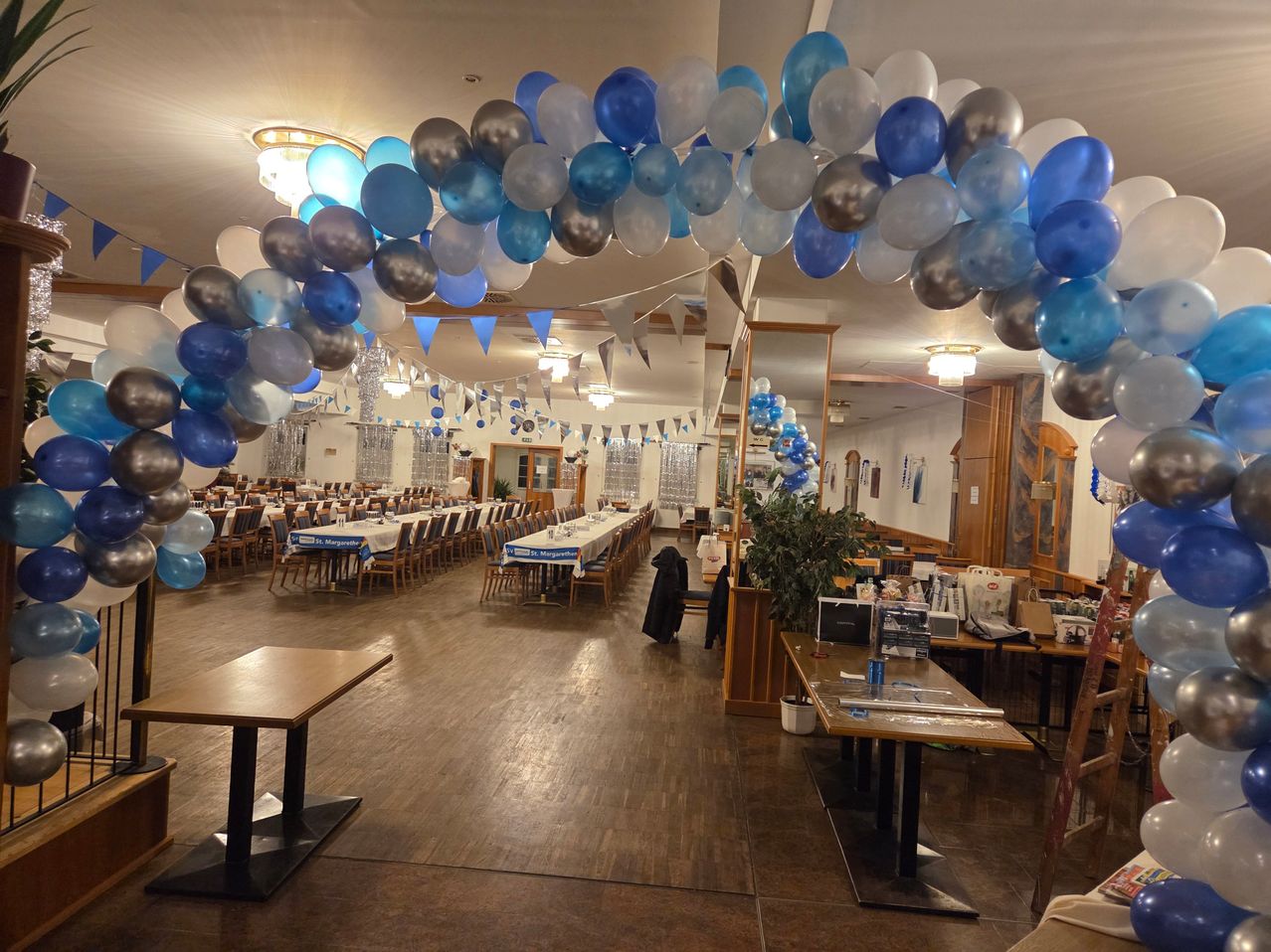 Ein Essbereich, der für eine besondere Veranstaltung dekoriert ist, mit Tischen und Stühlen, einer Decke, die mit blauen und weißen Luftballons geschmückt ist, und einem Spiegel an der Wand.