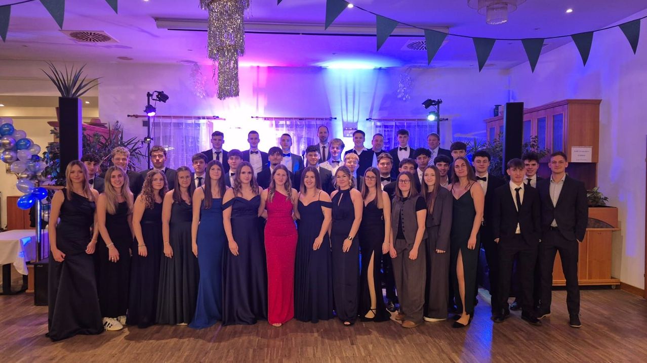 Eine Gruppe junger Menschen in formeller Kleidung posiert für ein Foto in einem Raum mit lila Beleuchtung und einem Holzboden.