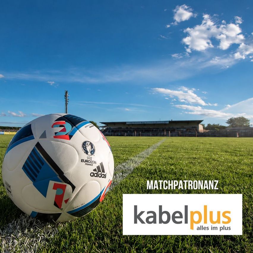 Ein Fußball mit Adidas-Logo steht auf einem Fußballplatz. Der Ball hat ein blaues und weißes Design mit roten Akzenten. Eine weiße Linie und ein Gebäude sind im Hintergrund sichtbar.