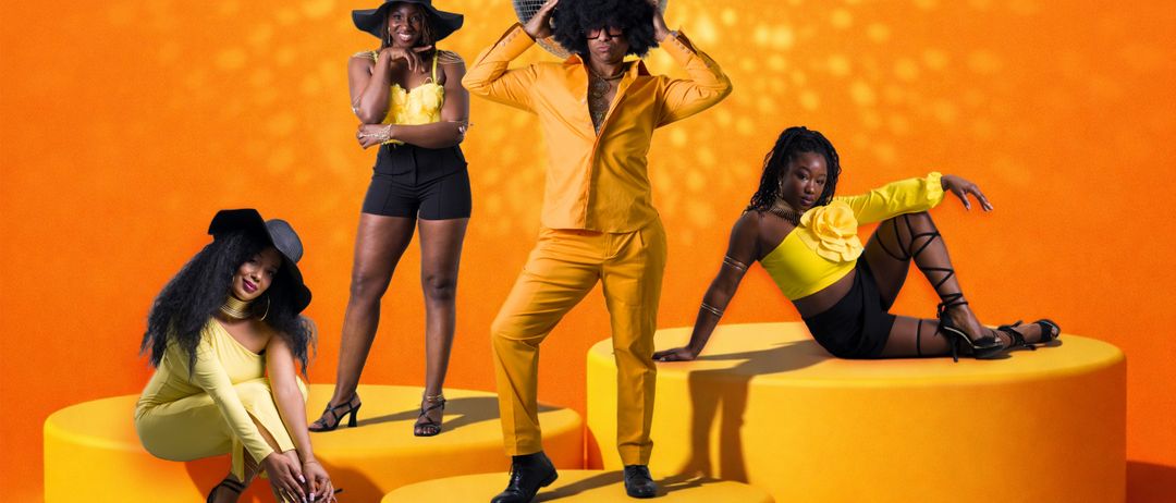 Three women and a man in yellow suits pose against an orange background, one holding a disco ball.