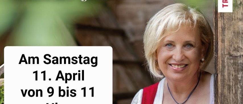 Eine lächelnde Frau in einem rot-weißen Kleid steht neben einem Holzpfosten mit Text zu einem April-Event.