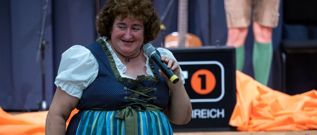 Eine Frau in einem traditionellen blauen Kleid mit weißen Punkten und einem grünen Gürtel hält ein Mikrofon und lächelt, möglicherweise auf der Bühne. Hinter ihr steht eine Gitarre und ein Schild mit der Nummer 1.