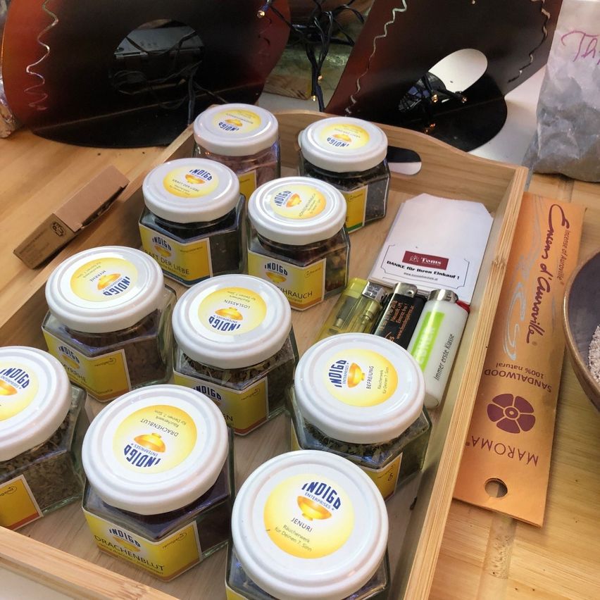 Ein Holztablett mit verschiedenen Gläsern von Indigo-Produkten, jeweils mit weißen Deckeln und gelben Etiketten. Zusätzliche Gegenstände umfassen ein Feuerzeug, ein kleines Büchlein und eine orangefarbene Verpackung.