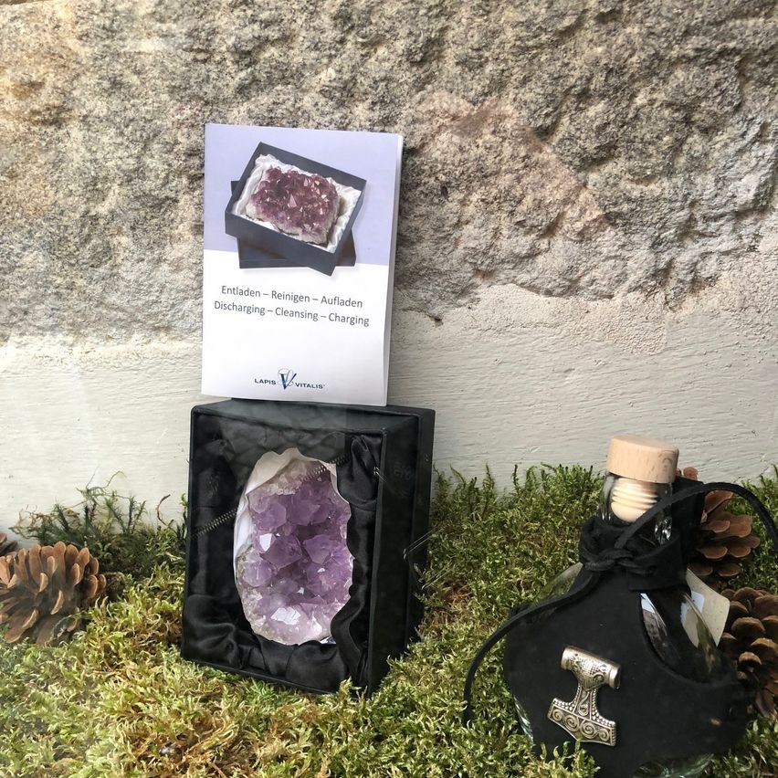 Eine schwarze Schachtel mit einem violetten Amethyst darin steht auf dem Boden, neben einer Glasflasche mit Korkenverschluss.