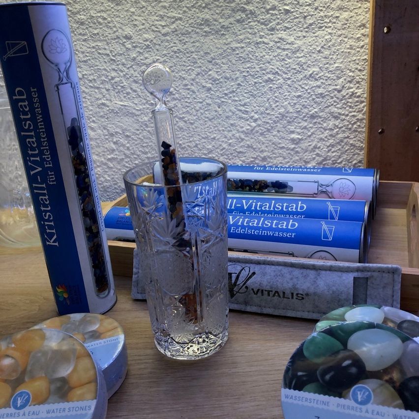 Ein Glas mit einem mit Steinen gefüllten Rohr auf einem Holztisch. Daneben eine Glasflasche mit einem Glasstab. Dahinter eine weiße Wand und mehrere blaue Schachteln mit der Aufschrift "Edelsteinwasser".