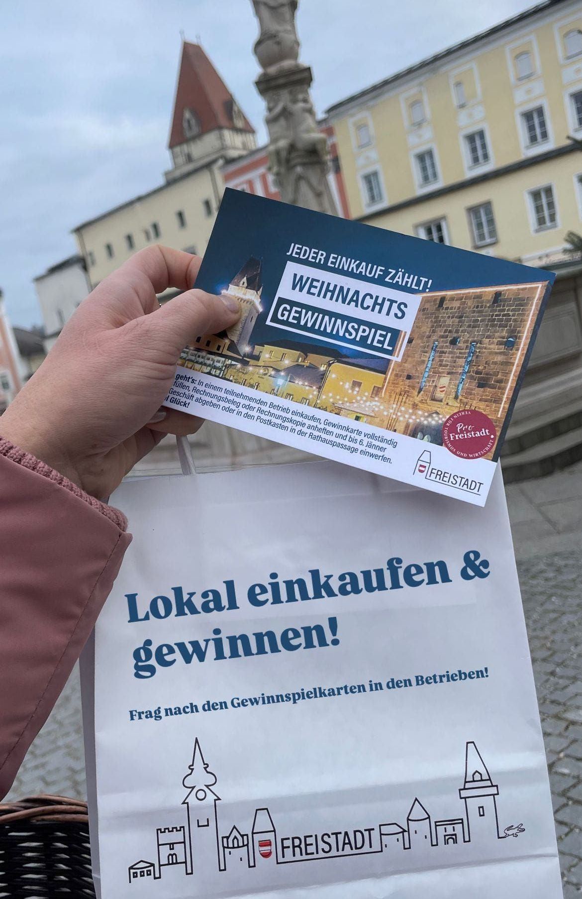 Eine Person hält einen Flyer mit einer Weihnachtsverlosung, die eine festliche Stadtansicht zeigt. Der Flyer sagt: 'Jeder Einkauf zählt! Weihnachtsgewinnspiel.' Der weiße Beutel, den sie hält, sagt: 'Lokal einkaufen und gewinnen!' Der Hintergrund zeigt eine Stadt mit Gebäuden und einem Turm.