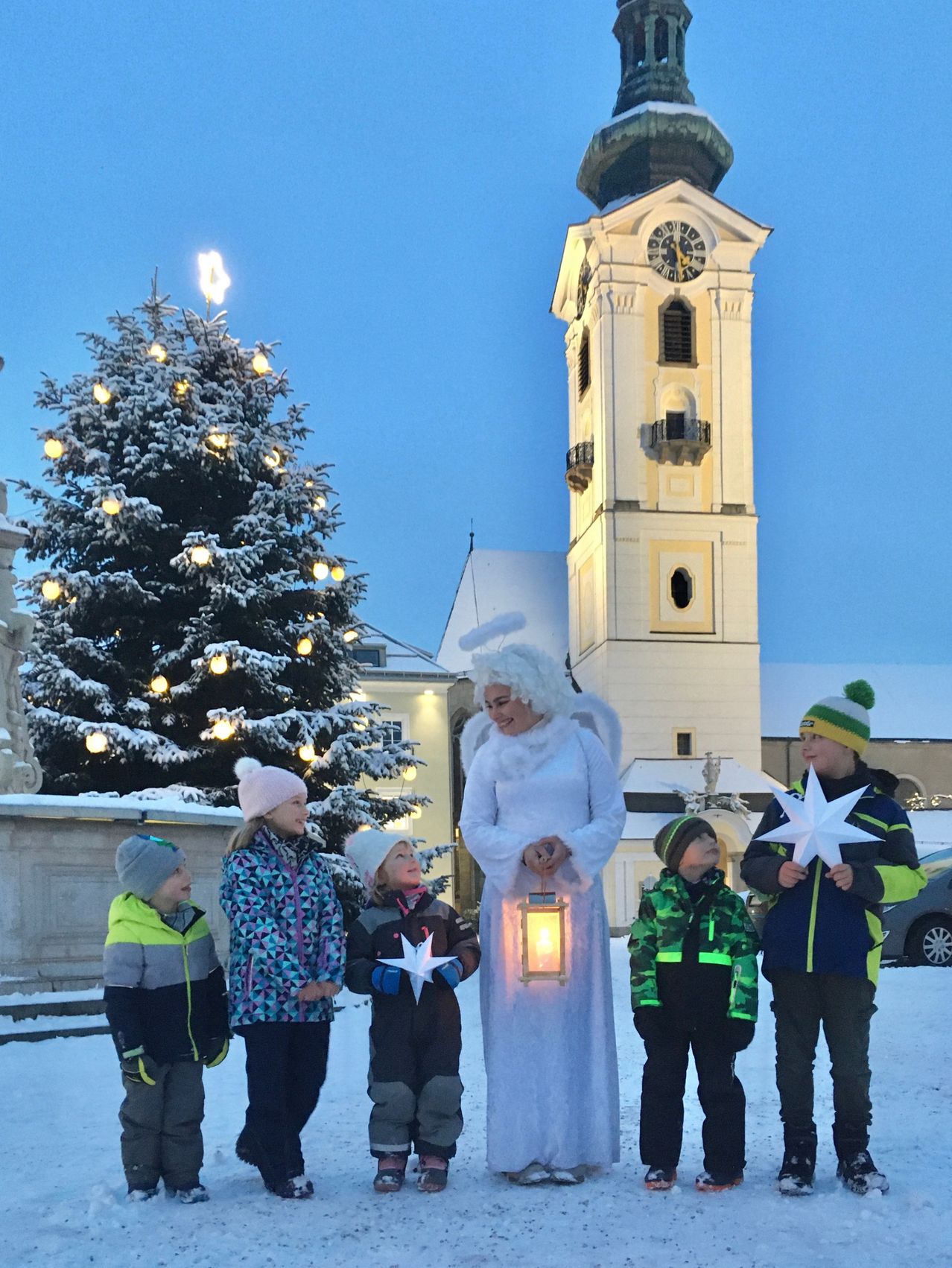 Eine Frau, die als Engel gekleidet ist, steht vor einer Gruppe von Kindern in Winterkleidung. Sie halten Papiersterne in der Hand und stehen vor einem Turm und einem Weihnachtsbaum.