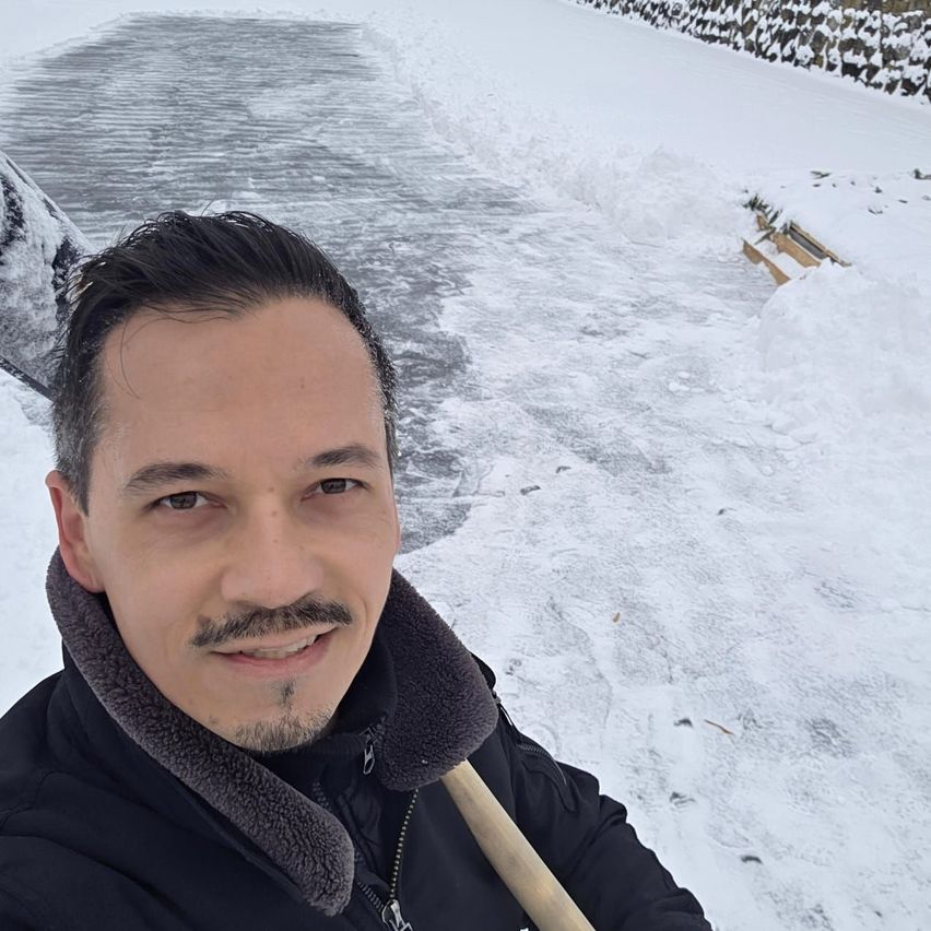 Ein Mann lächelt für ein Selfie mit einer Schaufel und steht in einer verschneiten Landschaft mit einem schmelzenden Eisweg.