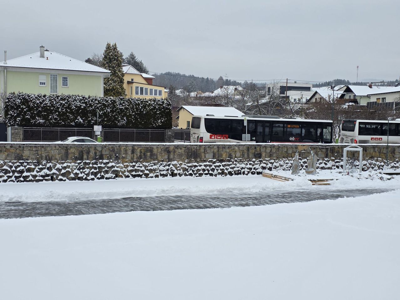Eine verschneite Landschaft mit zwei Bussen, die an einer Steinmauer geparkt sind, und einem Gebäude mit schneebedecktem Dach.