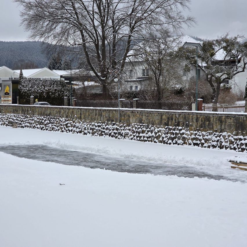 Eine schneebedeckte Landschaft mit einer Steinmauer im Vordergrund, durch die ein teilweise geschmolzener Fluss fließt. Bäume und Häuser, die mit Schnee bedeckt sind, befinden sich im Hintergrund, zusammen mit einem bewölkten Himmel.