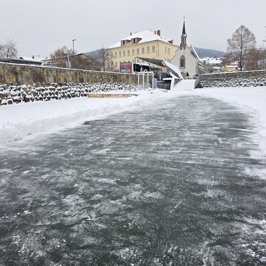 Eine verschneite Szene mit einem zugefrorenen Eisfeld vor einer Steinmauer, einem Gebäude mit einem Turm und einer Person, die in der Nähe einer Kirche geht.