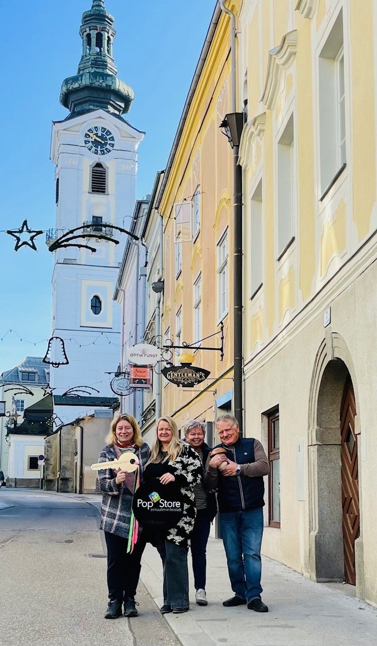 Vier Personen posieren für ein Foto vor einem Gebäude. Sie stehen in der Nähe eines Turms mit einer Uhr und einem dekorativen Stern.