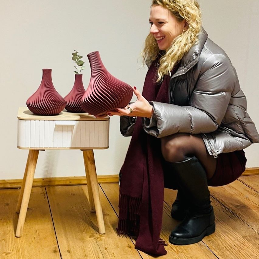 Eine Frau kniet auf einem Holzboden und hält eine große, dunkelrote Vase. Sie lächelt und scheint die Vase zu betrachten. Hinter ihr steht eine kleinere, dunkelrote Vase auf einem Tisch.