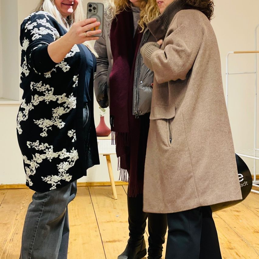 Drei Frauen stehen in einem Raum, eine macht ein Selfie. Die Frau auf der linken Seite trägt einen Blumenpullover, die mittlere Frau einen braunroten Schal und die rechte Frau einen beigen Mantel. Dahinter steht ein Tisch mit einer Vase.