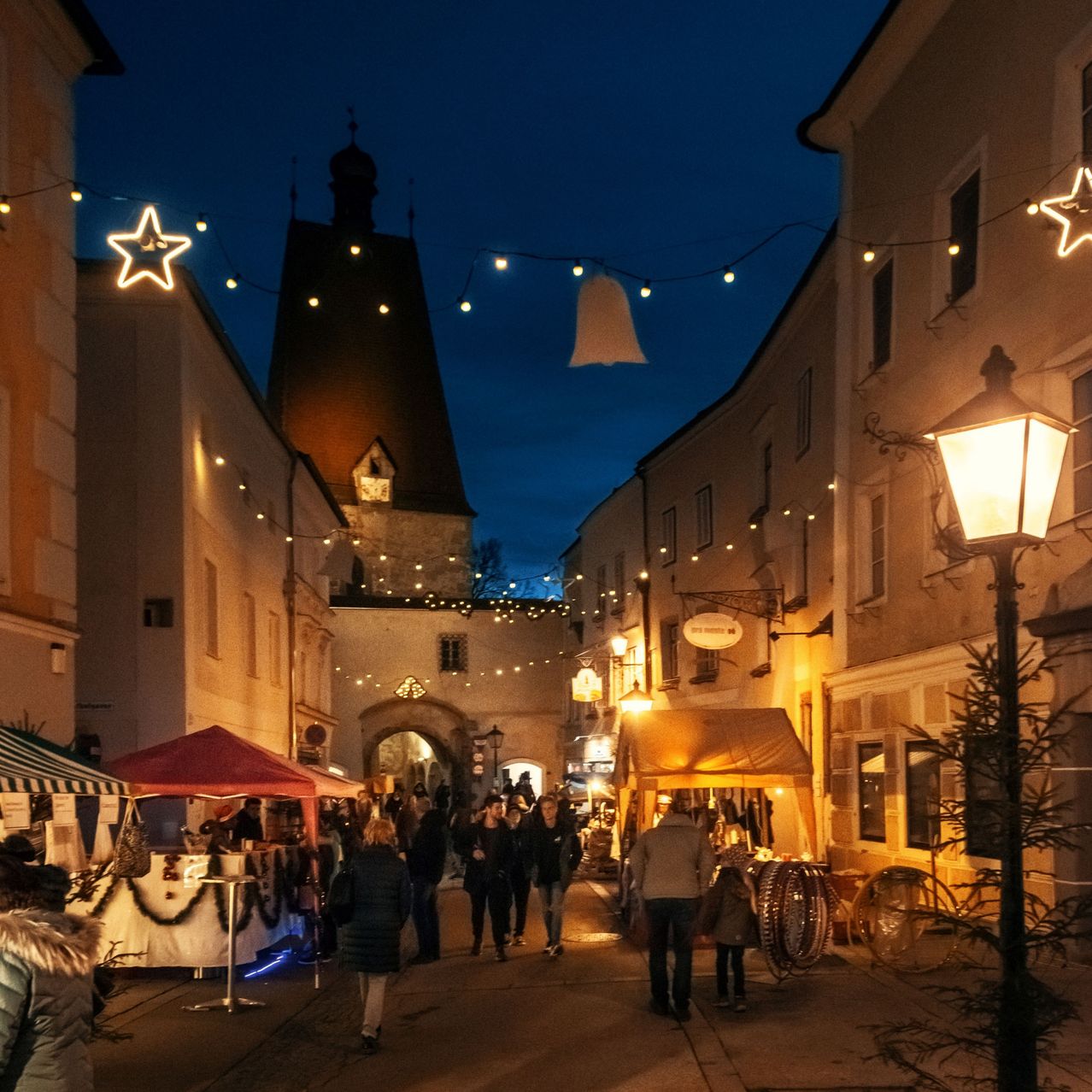 Ein Straßenmarkt bei Nacht ist voller Menschen. Stände sind auf beiden Seiten der Straße aufgebaut, mit Dekorationen und Lichtern, die den Bereich erhellen.
