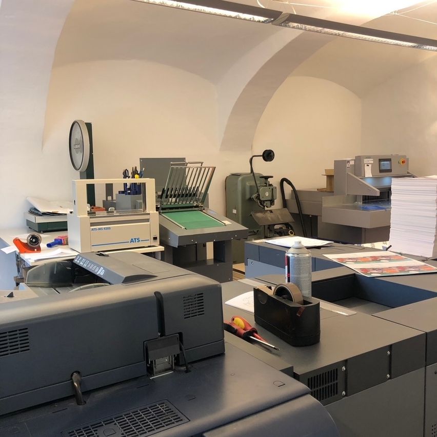 A printing room with various machines and tools, including a large printer in the foreground, other printers, and a machine with a clock and scissors.