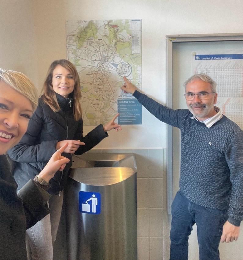 Three people stand by a trash bin, pointing at a map on the wall with a smiling man pointing at a map on the wall.