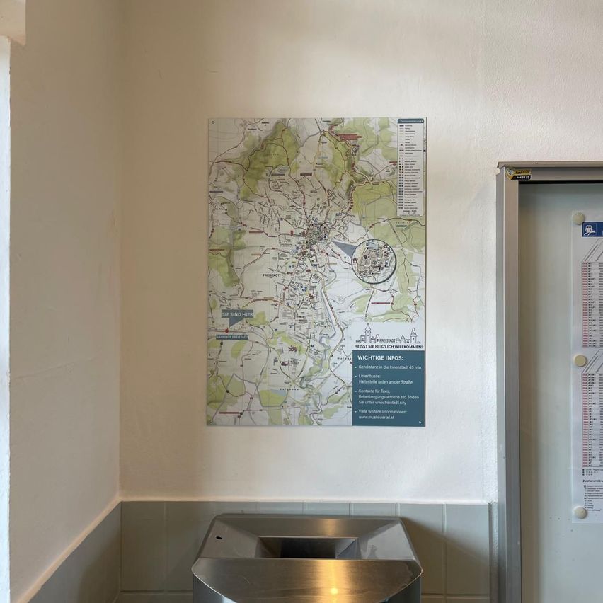 A map with 'You are here' sign is mounted on a wall, with a metal trash bin below and a white door on the right.