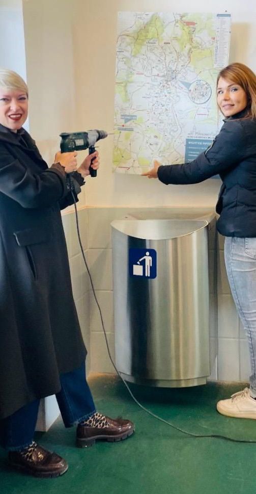 Two women are standing in a room, one holding a drill and the other pointing at a trash bin. They are both smiling. A map is pinned on the wall behind them.