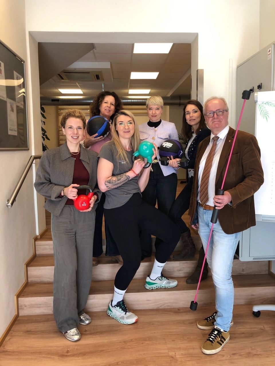Eine Gruppe von Menschen in Trainingskleidung posiert für ein Foto in einer Treppe, hält Kettlebells und lächelt.