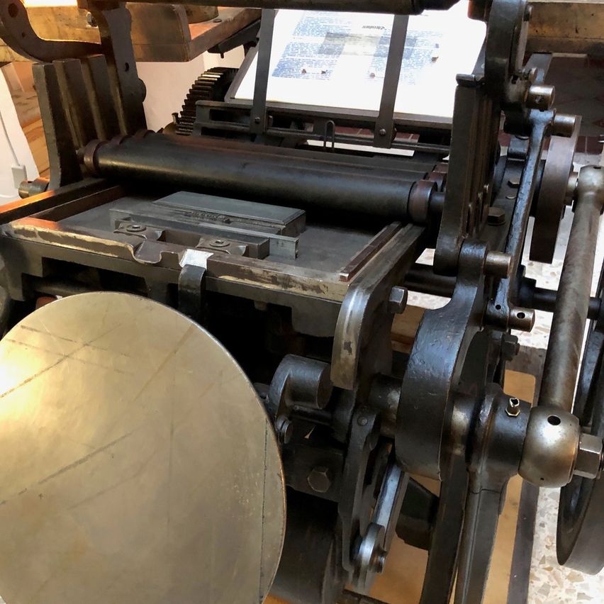 Eine alte Druckmaschine mit metallenem Rahmen, Zahnrädern und einem großen Metallrad wird drinnen ausgestellt. Ein Glasrahmen befindet sich auf der Maschine.