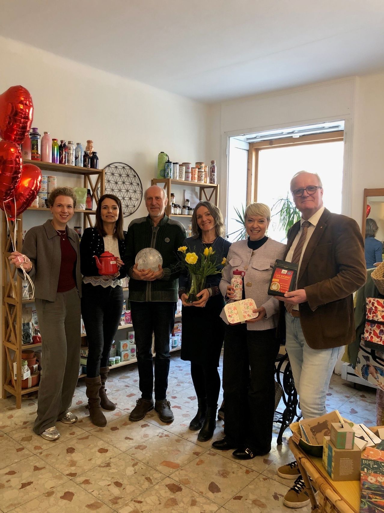Eine Gruppe von Menschen steht in einem Raum, hält Geschenke und Blumen und hat Regale und Ballons im Hintergrund.