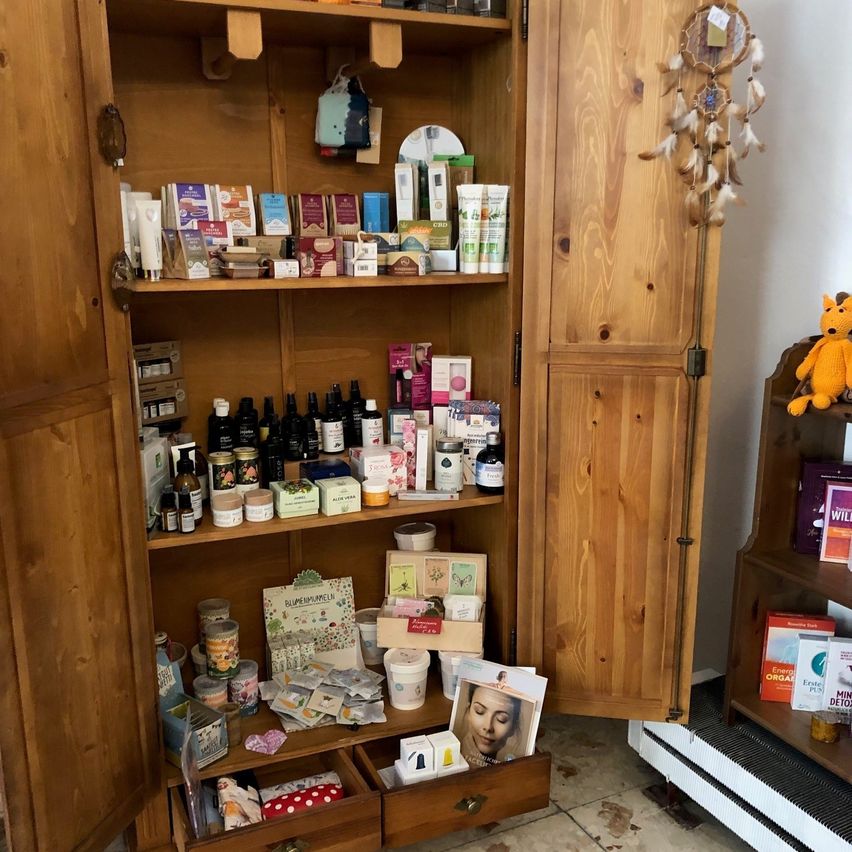 Ein hölzerner Schrank mit mehreren Regalen, gefüllt mit verschiedenen Produkten wie Flaschen, Schachteln und Gläsern. Ein Traumfänger hängt an der Tür.