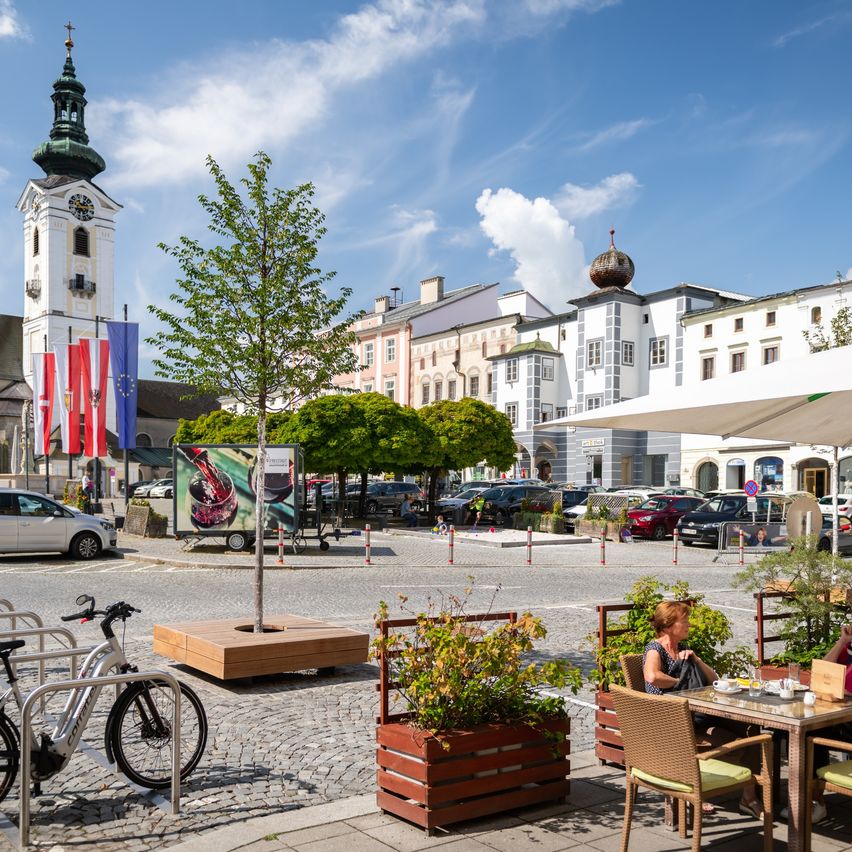 Ein Stadtplatz mit einem Glockenturm, geparkten Autos und einer Frau, die an einem Tisch im Freien unter einem Schirm sitzt.