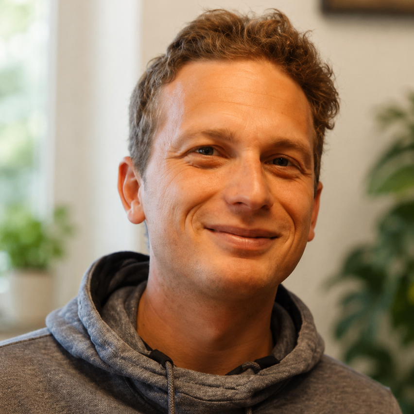 Ein Mann mit lockigem Haar trägt eine graue Kapuzenjacke und lächelt in die Kamera. Er steht vor einer weißen Wand mit einer Topfpflanze im Hintergrund.