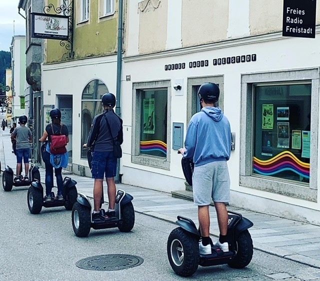 Vier Personen fahren auf einem Segway auf einer Stadtstraße, wobei ein Gebäude im Hintergrund ein Schild mit der Aufschrift 'Freies Radio Freistadt' zeigt.
