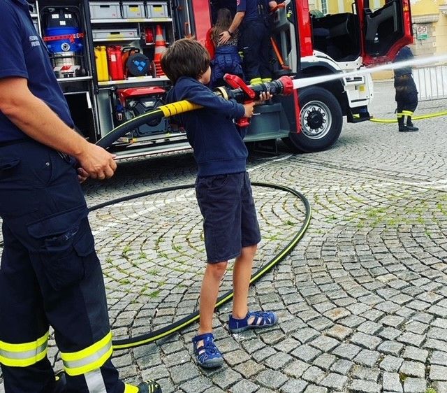 Ein junger Junge hält eine Feuerwehrspritze und sprüht Wasser, während ein Feuerwehrmann neben ihm steht.