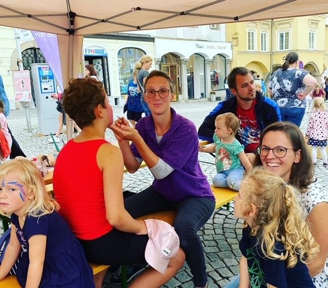 Eine Gruppe von Menschen sitzt auf einer Bank unter einem Zelt. Eine Frau schminkt ein Mädchen. Eine andere Frau hält ein Baby. Hinter ihnen sind Gebäude und Menschen, die auf der Straße gehen.