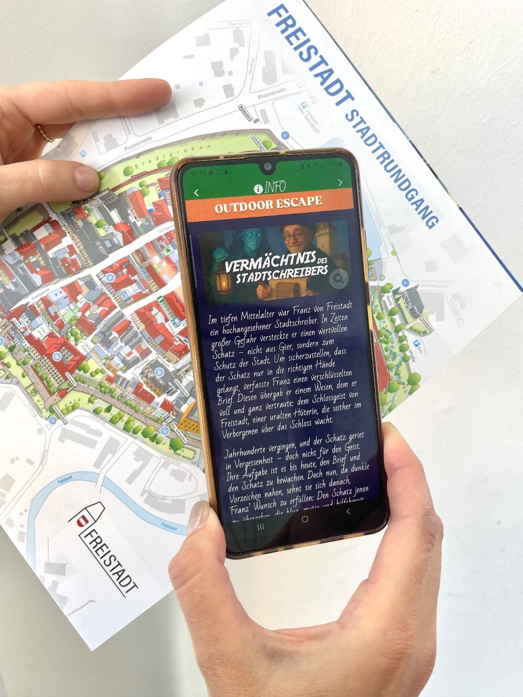 Eine Person hält ein Smartphone mit einer Outdoor-Escape-Game-App, auf der eine Karte von Freistadt im Hintergrund zu sehen ist.