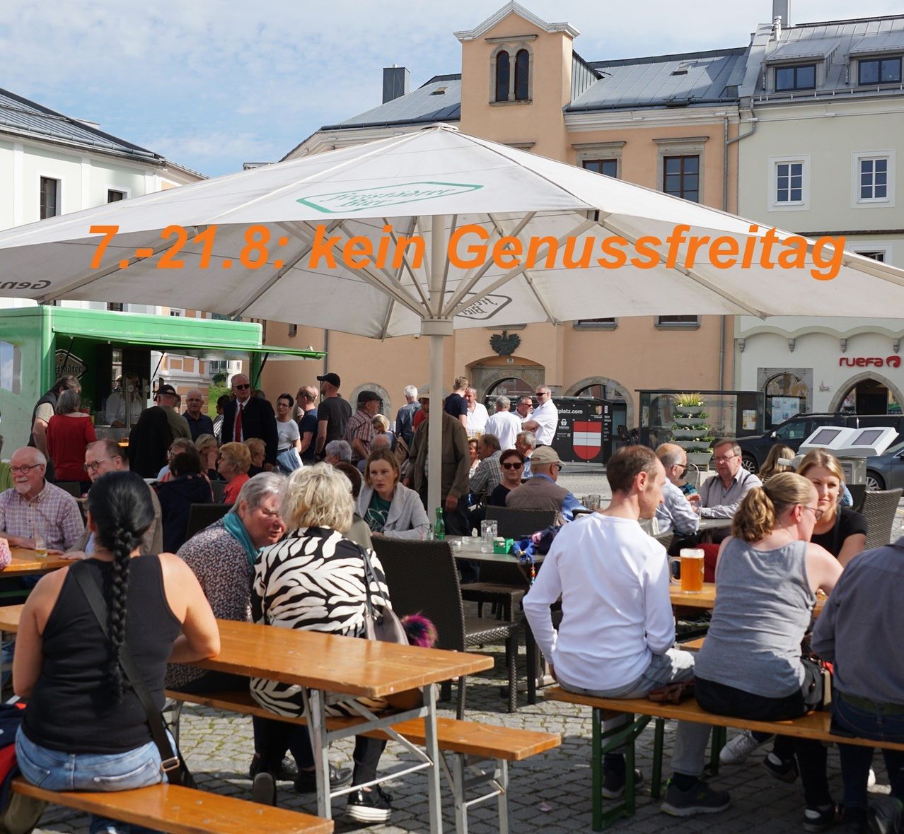 A bustling outdoor cafe with many people seated under a large umbrella. The cafe is surrounded by buildings, and there is a food truck parked nearby. The image has a watermark with the text '7-21.8: kein Genussfreitag'.