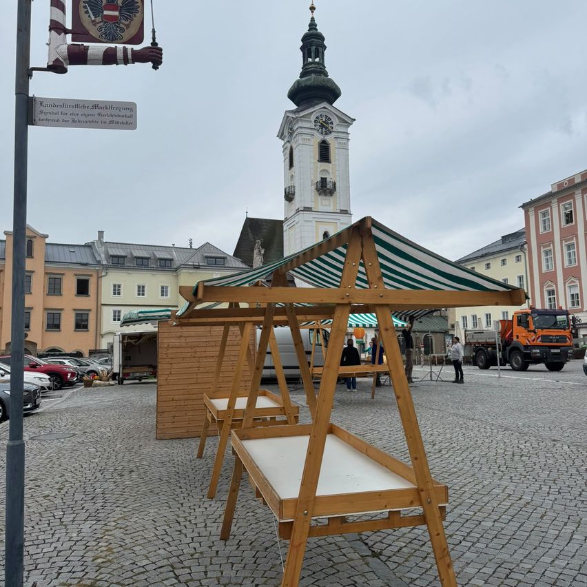 Ein hölzerner Marktstand mit einem grün-weiß gestreiften Baldachin steht auf einem Kopfsteinpflasterplatz, mit einem Glockenturm und Gebäuden im Hintergrund.