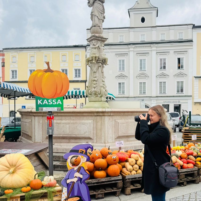 Eine Frau fotografiert einen Kürbis-Stand vor einem Brunnen und einem historischen Gebäude.