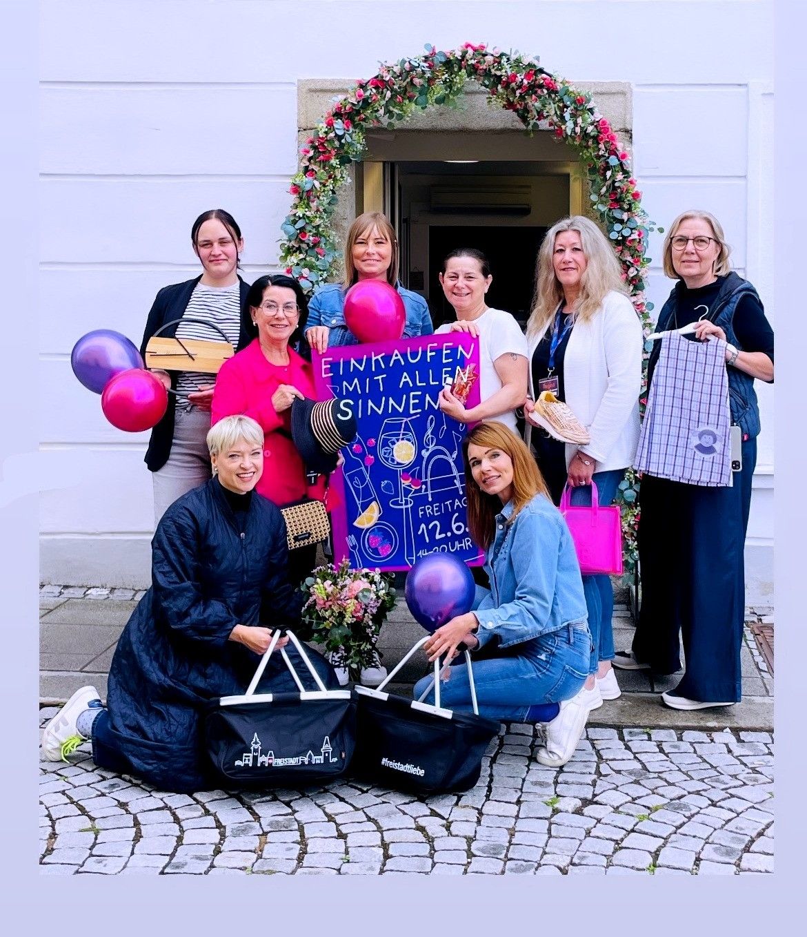 Eine Gruppe von Frauen mit Ballons und Taschen posiert vor einem Gebäude mit einem Blumenbogen und einem Banner mit der Aufschrift 'Einkaufen mit allen Sinnen'.
