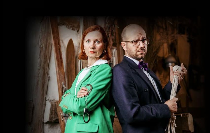 Ein Mann und eine Frau stehen zusammen, schick gekleidet. Die Frau hält ein Paar Handschellen. Der Mann hält eine Seil und sieht ernst aus. Sie scheinen sich in einem Gebäude zu befinden.