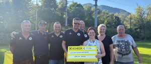 Eine Gruppe von Menschen steht lächelnd auf einem Feld, eine Frau hält einen großen gelben Scheck. Dahinter ein gelbes Banner und eine landschaftliche Aussicht mit Bäumen und Bergen.