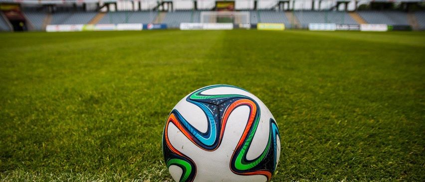 Ein Fußball mit bunten Designs liegt auf einem grünen Rasen. Der Hintergrund ist unscharf mit einem leeren Stadion in der Ferne.