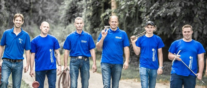 Fünf Männer in blauen Poloshirts stehen in einer Reihe auf einem Pfad, halten Werkzeuge und lächeln für die Kamera. Der Mann auf der rechten Seite hält eine Schaufel und der Mann in der Mitte hält ein Metallobjekt.
