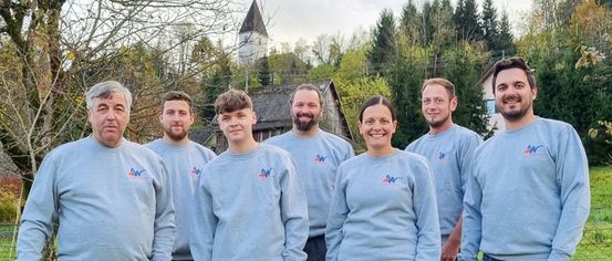 Eine Gruppe von sieben Erwachsenen, drei Männer und vier Frauen, tragen blaue Sweatshirts mit einem Logo und stehen auf einem Feld mit Bäumen und einem Haus im Hintergrund.