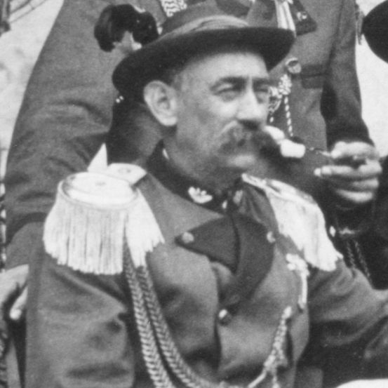 Ein historisches Bild zeigt einen älteren Mann in Uniform, der eine Pfeife raucht. Er trägt einen Hut und Epauletten. Ein anderer Soldat steht hinter ihm.